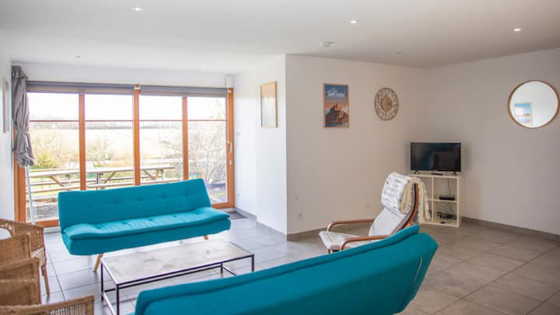 Salon lumineux du gîte Mistral avec canapés turquoise et baies vitrées sur la campagne bretonne près de Saint-Malo