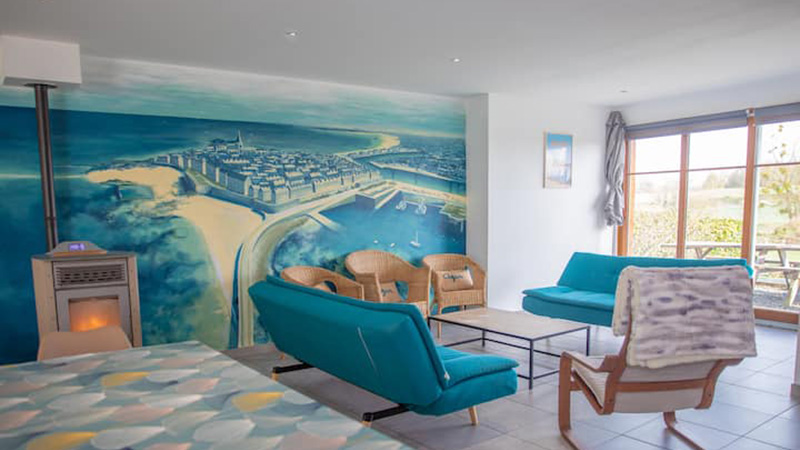 Salon du gîte Mistral avec grande fresque murale de Saint-Malo intra-muros, poêle à granulés et canapés turquoise